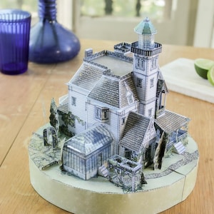 Op de afbeelding: Een driedimensionaal papieren model van een huis met meerdere verdiepingen, een vuurtoren, een glazen serre en een tuinpad, gemonteerd op een ronde basis. De structuur heeft witte gevels, grijze daken met dakpannen en groene beplanting.
