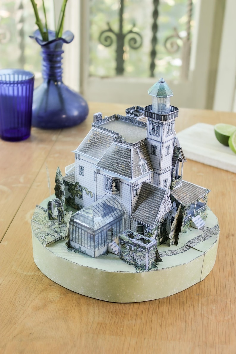 Op de afbeelding: Een driedimensionaal papieren model van een huis met meerdere verdiepingen, een vuurtoren, een glazen serre en een tuinpad, gemonteerd op een ronde basis. De structuur heeft witte gevels, grijze daken met dakpannen en groene beplanting.