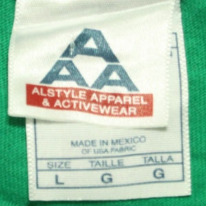 May include: White tag with a blue triangle logo that says "AA Alstyle Apparel & Activewear" and "Made in Mexico of USA Fabric". The tag also shows the size in L, Taille in G, and Talla in G.