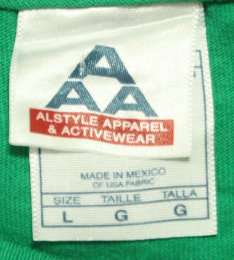 May include: White tag with a blue triangle logo that says "AA Alstyle Apparel & Activewear" and "Made in Mexico of USA Fabric". The tag also shows the size in L, Taille in G, and Talla in G.