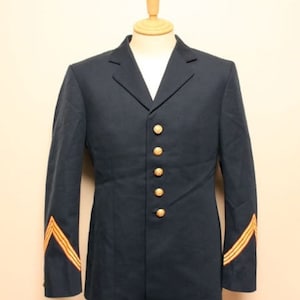 Puede incluir: Una chaqueta azul oscuro con botones dorados y rayas de galón doradas en las mangas. La chaqueta está en un maniquí.