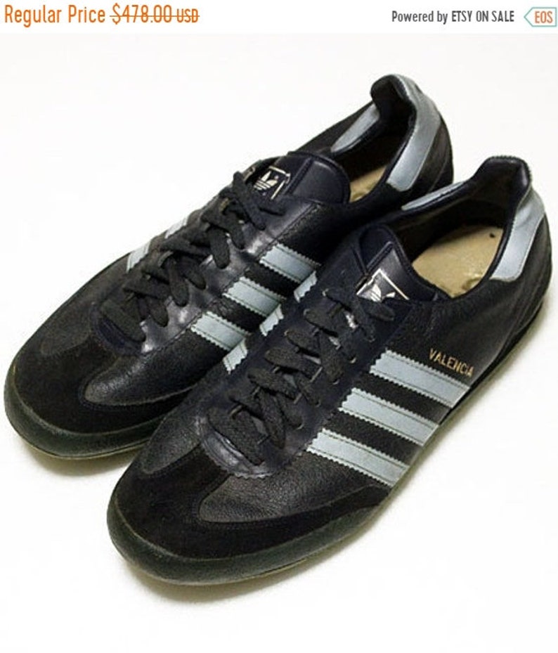 以下が含まれることがあります： 白と銀のストライプが入った黒の革製のadidasのスニーカーのペア。靴はスエードのつま先で、側面に「バレンシア」という文字が印刷されています。