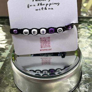 May include: A black beaded bracelet with purple letter beads spelling out the word "WEIRD". The bracelet is on a white card with the text "Thank you for shopping with us" and a QR code with the text "WitchyDelight".