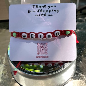 Può includere: Un braccialetto di corda rosso con perline bianche che compongono la parola "WEIRD" e perline verdi alle estremità. Il braccialetto è su un cartoncino bianco con il testo "Grazie per aver acquistato da noi" e un codice QR con il testo "WitchyDelight".