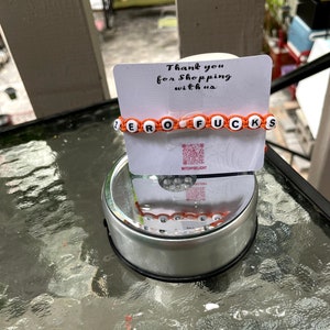 May include: Orange and white beaded bracelet with the words "ZERO F*CKS" spelled out. The bracelet is on a white card with the text "Thank you for shopping with us" and a QR code.