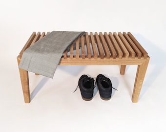 May include: A wooden bench with a slatted top and four legs. A gray blanket is draped over the bench and two black shoes are on the floor in front of it.