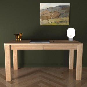 Könnte beinhalten: Ein heller Eichenschreibtisch mit zwei Schubladen, einem Laptop, einer kleinen goldenen Katzenfigur und einer modernen weißen Lampe. Ein Landschaftsbild hängt über dem Schreibtisch. Der Schreibtisch steht vor einer dunkelgrünen Wand.