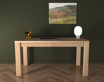 May include: A light oak wood desk with four sturdy legs and two front drawers. On the desk surface sits a closed dark gray laptop, a small metallic gold cat figurine, and a white spherical lamp with a gray base. The desk has a smooth finish.