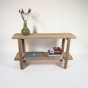 Peut inclure: Une console en bois clair dotée de deux étagères horizontales et de pieds inclinés. Sur l'étagère supérieure se trouve un vase en céramique verte avec des branches séchées. L'étagère inférieure contient un ordinateur portable argenté et quatre livres.