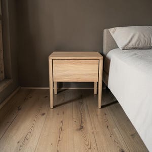 Op de afbeelding: Een lichthouten nachtkastje met een enkele lade met een gladde, platte voorkant en vier taps toelopende poten. Het meubel is gemaakt van natuurlijk hout met een zichtbare houtnerf en heeft een eenvoudig, rechthoekig ontwerp.