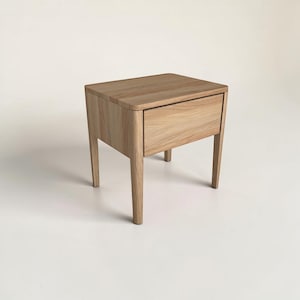 May include: A light-toned wooden nightstand featuring a rectangular top with rounded corners, a single front-facing drawer with a smooth grain finish, and four tapered legs that provide a stable base for this piece of bedroom furniture.