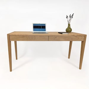 May include: A light oak desk with two drawers, a laptop, a small green vase with decorative stems, and a notebook. The desk has angled legs and a minimalist design, suitable for a home office or workspace.