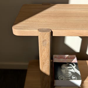 May include: A light-colored wooden side table featuring a smooth rectangular top and a vertical leg joined with a visible tenon. The table is positioned above a book titled 20th Century Photography, which features a black and white portrait.