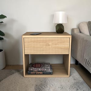 May include: A light wood nightstand with a single drawer and open shelf. A white lampshade sits atop a dark green geometric base. Two books are on the shelf, and a phone rests on the top. A potted plant is to the left.