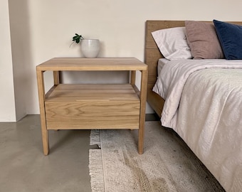 May include: A light-toned wooden nightstand featuring a flat top surface, an open shelf area, and a single drawer with a horizontal cutout handle. The nightstand is constructed with four straight legs and displays a visible natural wood grain pattern.