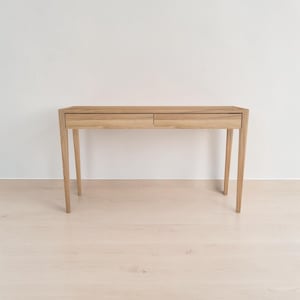 May include: A light brown wooden console table featuring a rectangular top and two side-by-side drawers with horizontal wood grain patterns. The table is supported by four tapered legs that angle outward, providing a stable base for the furniture piece.