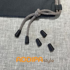 Pode incluir: Um saco de cordão cinzento-escuro com cordões tecidos cinzentos e pontas de plástico preto. Três pontas de plástico preto adicionais estão soltas por perto. O texto ADDIPA style está impresso em laranja e castanho na parte inferior.
