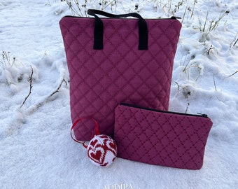 May include: A set of two quilted bags in a deep pink colour. The larger bag has two black handles and a zipper closure. The smaller bag is a flat pouch with a zipper closure. Both bags have a diamond quilted pattern. A small red and white knitted ornament is in the foreground.