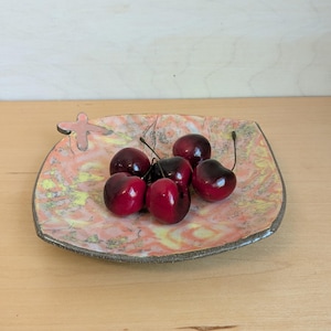 May include: A square ceramic dish with a pink and yellow marbled glaze. The dish has a small cross-shaped handle on one side. The dish is filled with red cherries.