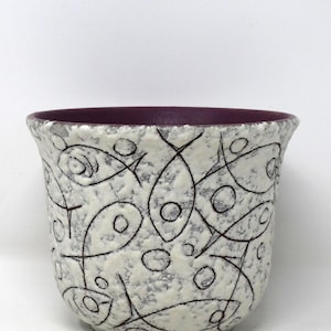 May include: A ceramic planter with a textured, off-white exterior adorned with a black fish-like pattern. The interior rim is a deep purple. This decorative pot is suitable for indoor plants.