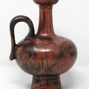 May include: A reddish-brown ceramic vase with a bulbous body, a handle, and a flared neck. The vase has a dark green accent on the rim and handle. The surface is marked with a network of dark lines.
