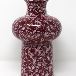May include: A cylindrical vase with a wide, rounded shoulder and a narrow neck, featuring a deep burgundy red base color with an irregular white speckled pattern covering the entire exterior surface of the vessel from the base to the rim.