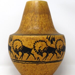 May include: A decorative, ceramic vase with a textured, yellow-brown finish. The vase features a black, repeating pattern of stylized rams around its midsection. The vase has a rounded base and a flared top.