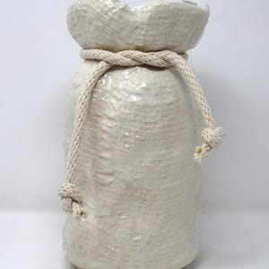 May include: Off-white ceramic vase with a textured surface, resembling a fabric sack. A thick, cream-colored rope is tied around the neck of the vase, with a knot on one side. The vase has a wide opening and a rounded top.