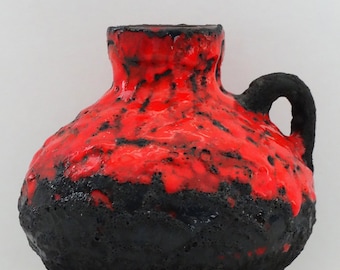 Puede incluir: Un jarrón de cerámica rojo y negro con una superficie texturizada. El jarrón tiene una base ancha y un cuello estrecho con una asa.