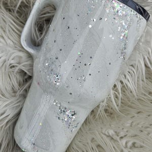 May include: White travel mug with a silver glitter design and a black lid. The mug has a handle and is sitting on a white fluffy surface.