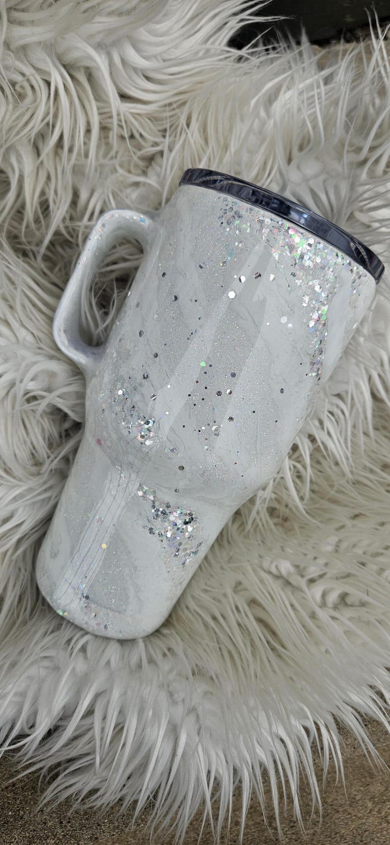 May include: White travel mug with a silver glitter design and a black lid. The mug has a handle and is sitting on a white fluffy surface.