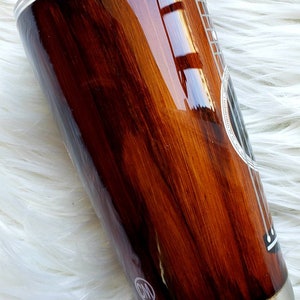 May include: A brown and black stainless steel tumbler with a wood grain design and a white guitar outline. The tumbler has a silver lid and a silver bottom.