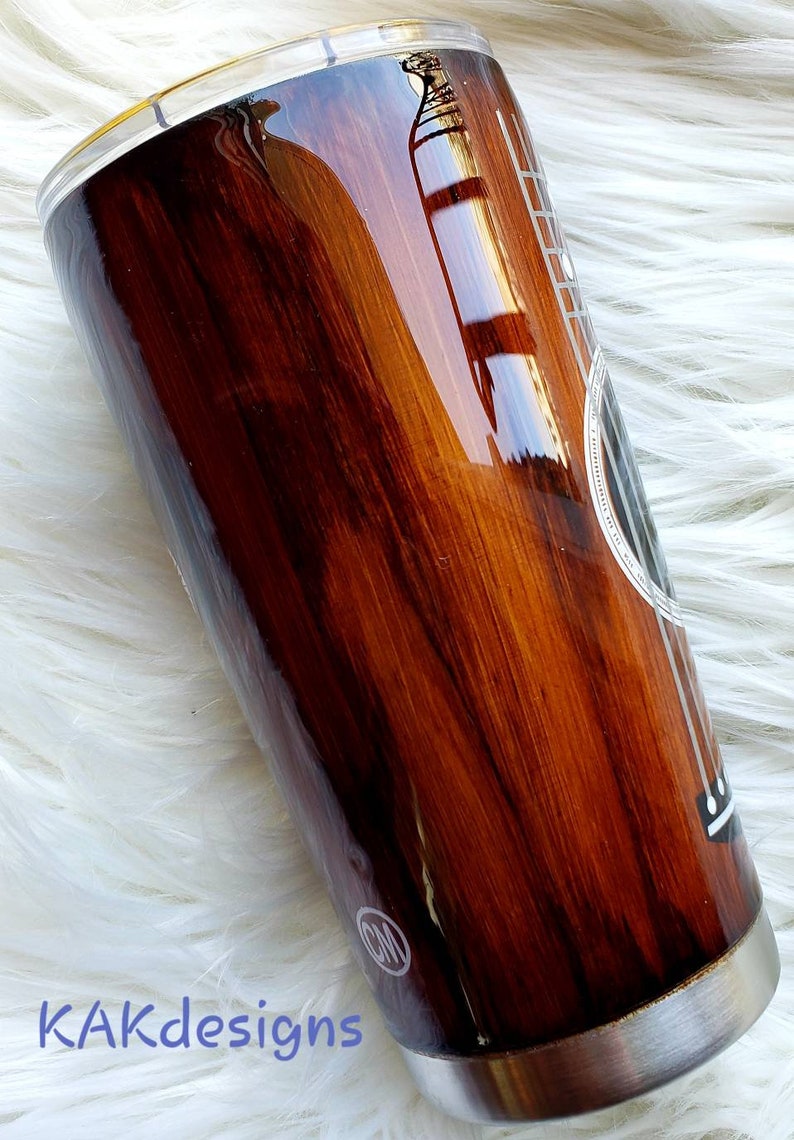 May include: A brown and black stainless steel tumbler with a wood grain design and a white guitar outline. The tumbler has a silver lid and a silver bottom.