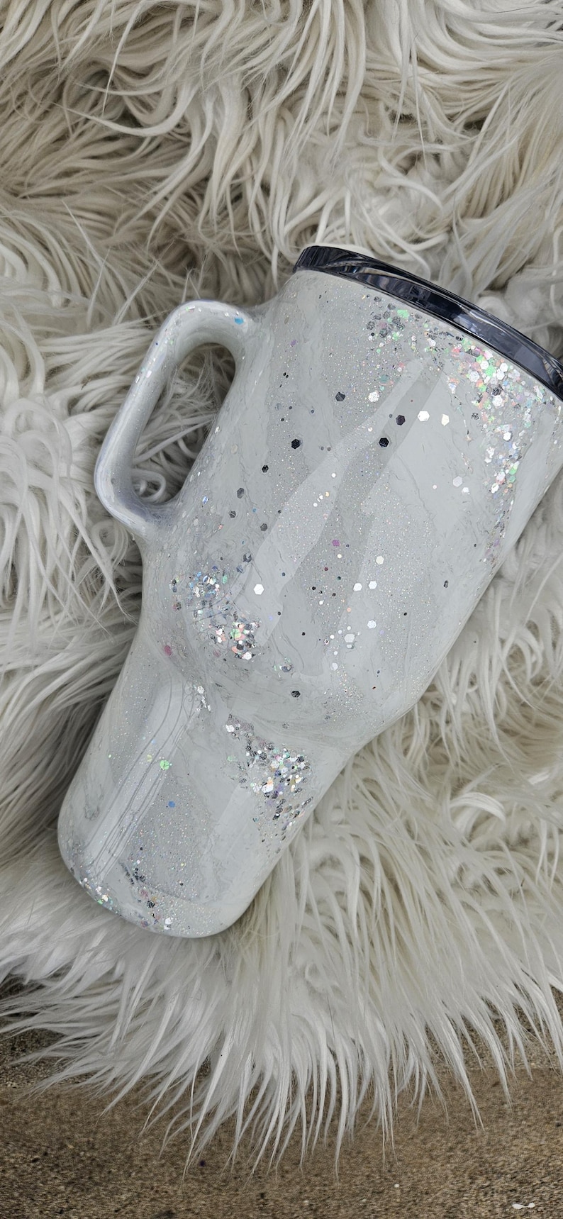 May include: White travel mug with a black lid and a handle. The mug is covered in silver glitter and has a marble-like design.