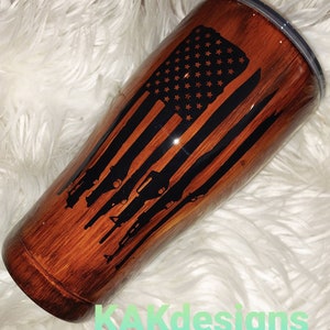 May include: A brown and black stainless steel tumbler with a wood grain design. The tumbler features a black silhouette of an American flag with guns in the stripes.