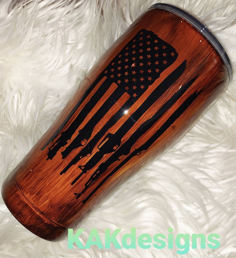 May include: A brown and black stainless steel tumbler with a wood grain design. The tumbler features a black silhouette of an American flag with guns in the stripes.