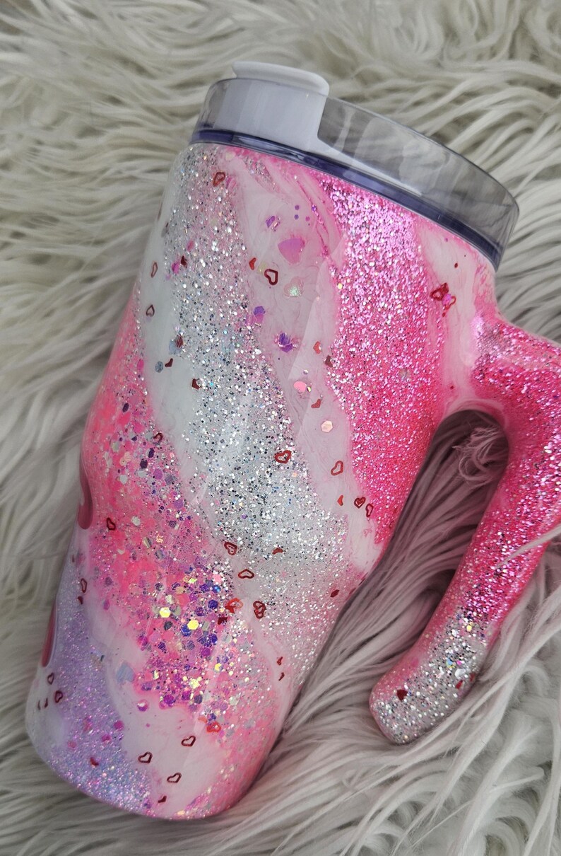 Puede incluir: Un vaso con purpurina y asa, con un dise&ntilde;o rosa, blanco y plateado. El vaso est&aacute; adornado con confeti en forma de coraz&oacute;n. La tapa es transparente con un deslizador blanco. Esta taza aislada est&aacute; dise&ntilde;ada para bebidas fr&iacute;as o calientes.