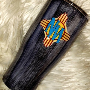 May include: A black and gray stainless steel tumbler with a wood grain design. The tumbler features a yellow, red, and blue graphic of the state of New Mexico with the letters "NM" in the center.