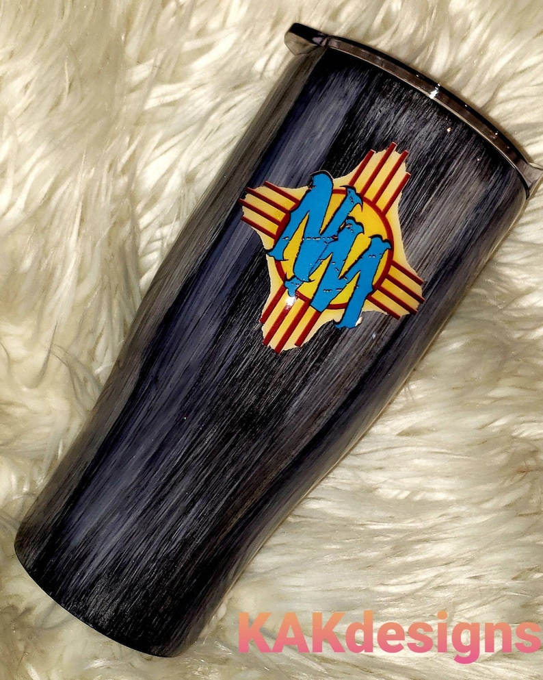 May include: A black and gray stainless steel tumbler with a wood grain design. The tumbler features a yellow, red, and blue graphic of the state of New Mexico with the letters "NM" in the center.