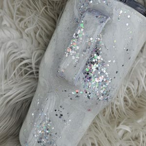 May include: A white tumbler with a silver glitter design and a black lid. The tumbler has a handle and is sitting on a white fluffy surface.