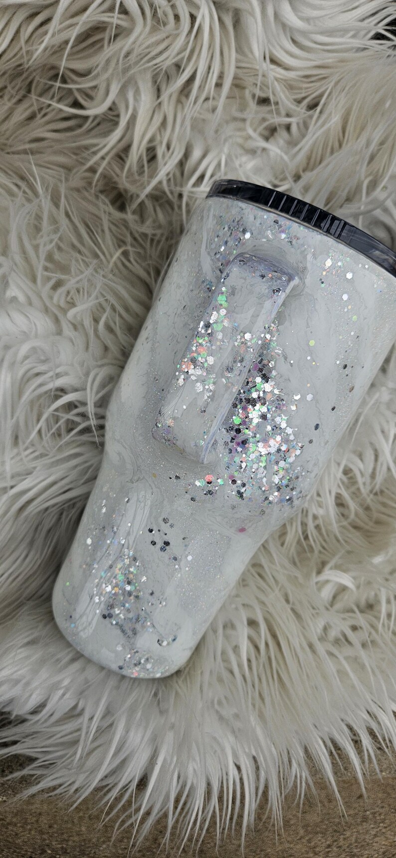 May include: A white tumbler with a silver glitter design and a black lid. The tumbler has a handle and is sitting on a white fluffy surface.