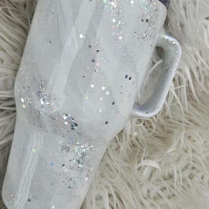 May include: A white travel mug with a black lid and a handle. The mug is covered in silver glitter.