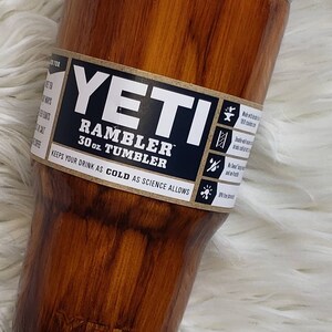 May include: A 30 ounce YETI Rambler Tumbler with a wood grain design. The tumbler is made of stainless steel and has a double-wall vacuum insulation to keep drinks cold. The label on the tumbler says "YETI Rambler 30 oz. Tumbler Keeps your drink as cold as science allows."