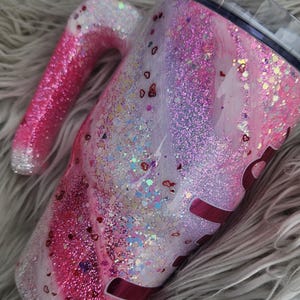 Puede incluir: Un vaso cubierto de purpurina con asa, con una mezcla de purpurina rosa, blanca y plateada. El dise&ntilde;o incluye confeti en forma de coraz&oacute;n y una tapa transparente. El asa es un degradado de rosa y plata.