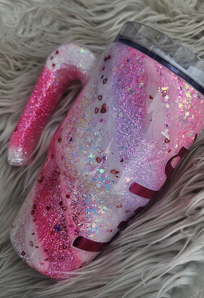 Puede incluir: Un vaso cubierto de purpurina con asa, con una mezcla de purpurina rosa, blanca y plateada. El dise&ntilde;o incluye confeti en forma de coraz&oacute;n y una tapa transparente. El asa es un degradado de rosa y plata.