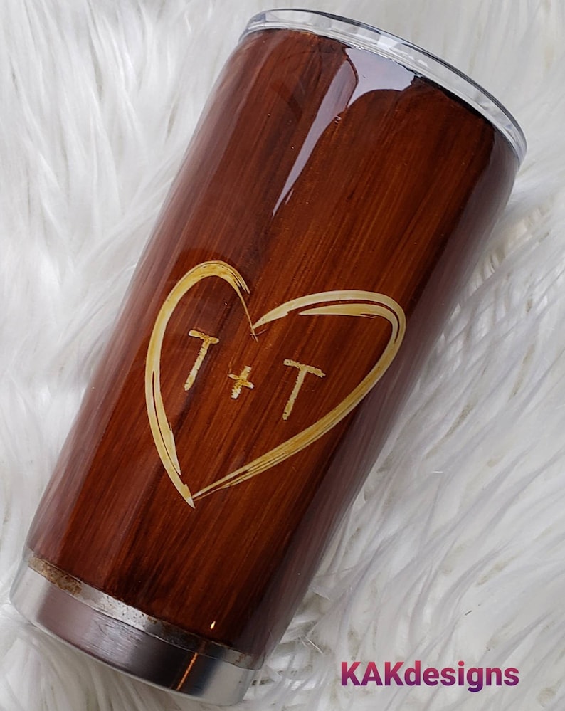 May include: A brown stainless steel tumbler with a wood grain design and a gold heart with the initials "T + T" engraved on it.