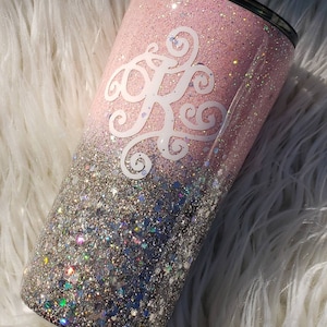 Puede incluir: Un vaso de glitter rosa y plateado con un monograma blanco. El vaso tiene un efecto degradado, con el glitter rosa que se funde en el glitter plateado.