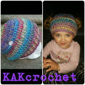 May include: A colorful crocheted hat with pigtails, featuring a variety of colors including pink, blue, green, and yellow. The hat is worn by a young child with a pink shirt and black pants. The image includes the text "KAKcrochet".