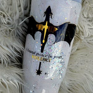 Könnte beinhalten: Ein weißer Becher mit einer schwarzen Drachen-Silhouette und goldenem Glitzer. Der Becher trägt den Text "I found peace in your violence".