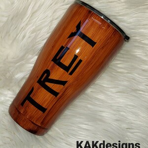 May include: A brown and black stainless steel tumbler with the name "TREY" printed on the side. The tumbler has a wood grain design.